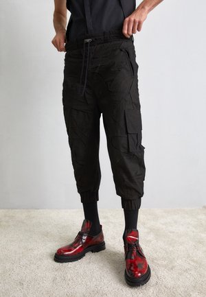 Homme portant un pantalon cargo court noir avec une taille à cordon de serrage et des chaussures à lacets rouge-noir brillantes, debout sur un tapis clair.