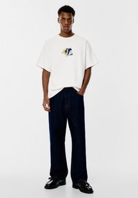 Weißes, übergroßes T-Shirt mit einem grafischen Druck auf der Brust, kombiniert mit dunkelblauen Jeans und schwarzen Schnürschuhen. Schlichtes und modernes Design.
