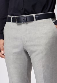 Grijze dressbroek met een gladde textuur, gecombineerd met een zwarte leren riem met een zilveren gesp. Het shirt is donker en met een kraag.