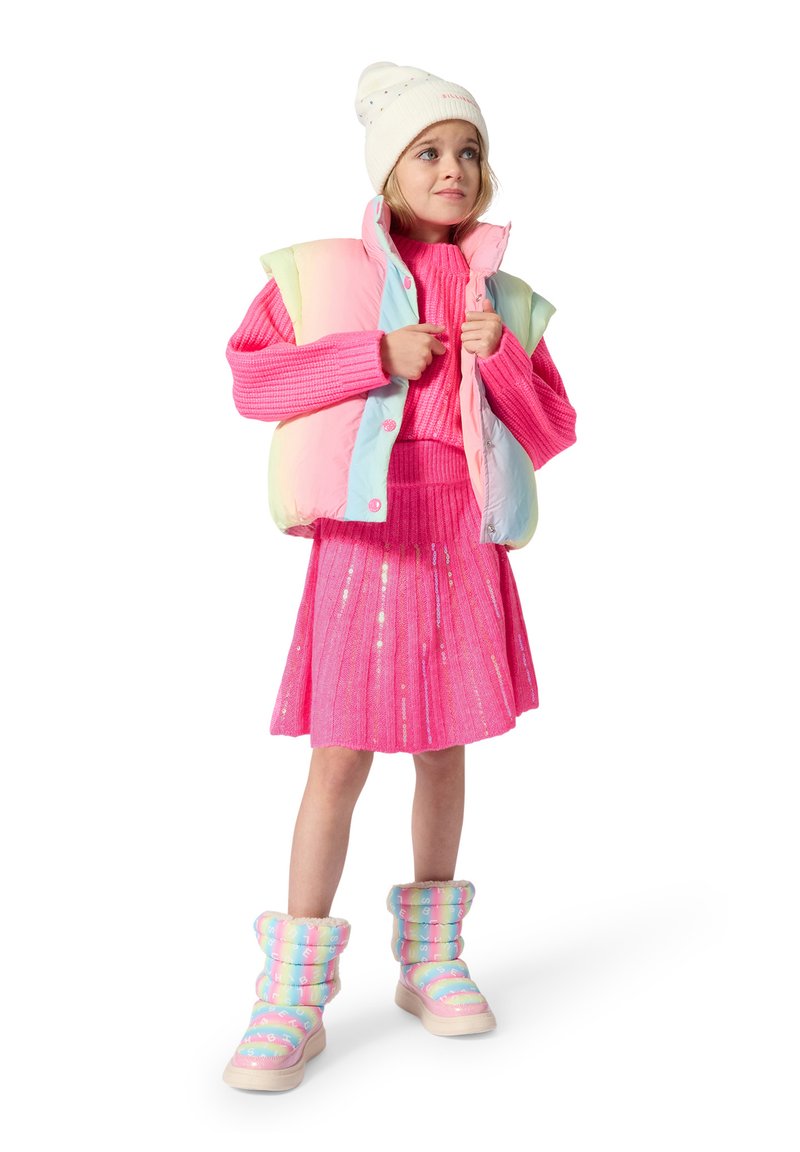 Jeune fille portant un bonnet en tricot blanc, un gilet arc-en-ciel pastel, un pull rose vif et une jupe, ainsi que des bottes d’hiver arc-en-ciel pastel, debout sur fond blanc.
