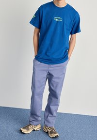 Modrá bavlněná tričko s žlutým logem Nike na hrudi, v kombinaci se světle fialovými kalhotami a béžovými teniskami s černými akcenty.