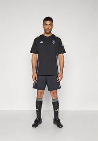 Camiseta deportiva negra con franjas verdes y amarillas, combinada con pantalones cortos a juego. Calcetas negras con franjas blancas y zapatos de fútbol negros.