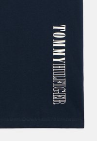 Mørkeblå bomulls-T-skjorte med vertikal hvit logo-print som leser "TOMMY HILFIGER" på venstre side. Enkelt design, korte ermer.