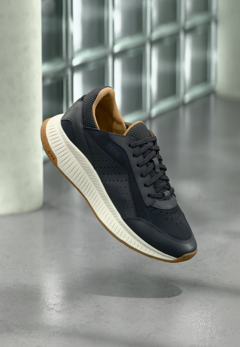 Must ja pruun vabaajajalanõu valge tallaga, paelte ja perforeeritud külgpaneelidega, näidatud hõljuvana halli põranda kohal.