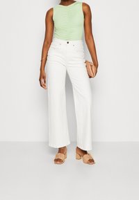 Body verde chiaro senza maniche con arricciature centrali, abbinato a jeans bianchi a gamba larga e sandali beige con tacco a blocco, mentre tiene una piccola borsa.