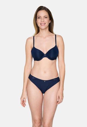 Frau mit glattem Haar, die einen marineblauen BH und passende, mit Spitze verzierte Höschen trägt, vor einem hellgrauen Hintergrund stehend und lächelnd.