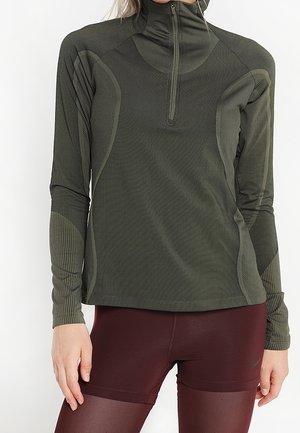 Frau trägt ein olivgrünes, langärmliges Sportoberteil mit Reißverschluss bis zum Viertel und bordeauxrote enganliegende Leggings, steht mit einer Hand entspannt neben dem Körper.