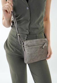 Borsa a tracolla grigia metallica con finitura testurizzata, chiusura superiore con zip, tasca frontale e tracolla regolabile, mostrata mentre viene tenuta accanto a una persona in un outfit verde.