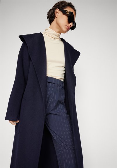 Cappotto di lana blu navy con ampie revers, abbinato a un dolcevita crema e pantaloni a righe. Occhiali da sole neri oversize completano il look.