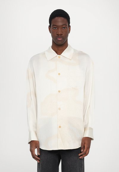 Camisa de botones color beige claro con un patrón sutil, mangas largas, collar puntiagudo y un bolsillo en el pecho. Presenta botones dorados claros.