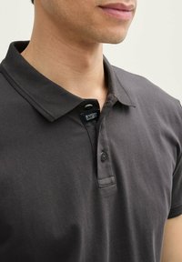 Polo shirt gris foncé avec un col, doté d'une patte de boutonnage à deux boutons et d'une étiquette en texte. Tissu lisse avec une texture subtile.