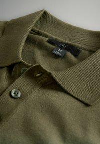 Olivgrünes Polohemd aus strukturiertem Stoff, mit klassischem Kragen, einer Knopfleiste mit drei Knöpfen und einem Etikett mit der Aufschrift "THE SET", Größe M.