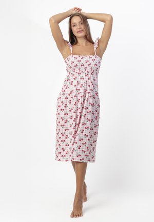 Mujer de pie descalza con los brazos levantados, vistiendo un vestido midi rosa con estampado de cerezas y tirantes con volantes en los hombros.