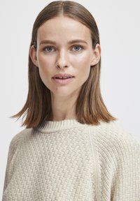 Pull en tricot de couleur beige clair avec un motif texturé et un col rond côtelé. Présente une coupe décontractée avec des coutures d'épaule bien définies.