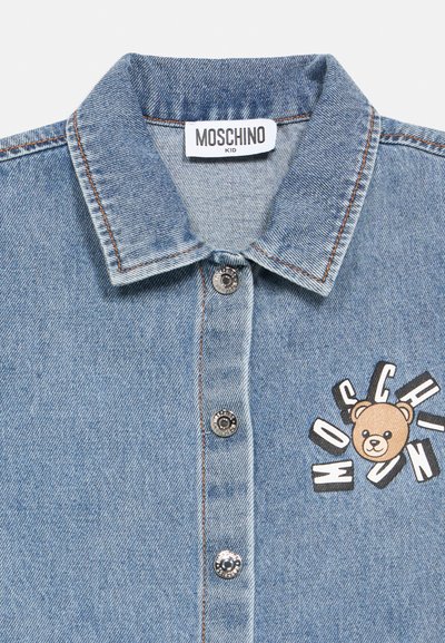 Πουκάμισο από denim με ανοιχτό γαλάζιο χρώμα, με μυτερό γιακά, ασημένια κουμπιά και γραφικό που απεικονίζει μια αρκούδα περιτριγυρισμένη από το κείμενο "MOSCHINO".