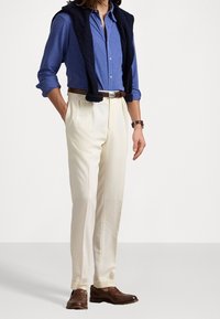 Blå knapp-up skjorta, krämfärgade byxor, marinblå kabelstickad tröja över axlarna, brun läderbälte och skor. Släta texturer och neutrala färger.