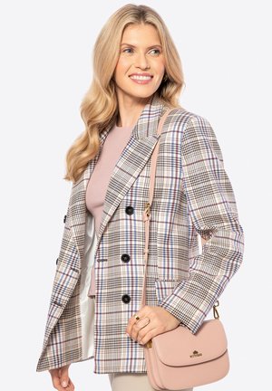 Mujer con cabello largo y rubio, vestida con un blazer a cuadros y una blusa rosa claro, sosteniendo un bolso de hombro rosa claro, sonriendo y mirando hacia la derecha.