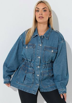 Blond vrouw die een blauwe spijkerjack draagt met grote zakken en knoopsluiting, gecombineerd met een zwarte broek, staande voor een effen achtergrond.