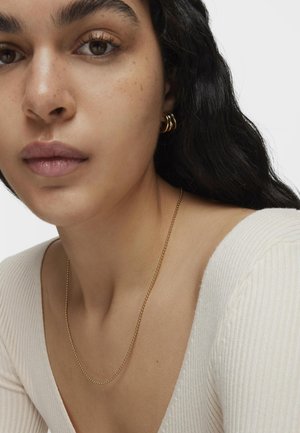 Collar de cadena de oro, textura delicada, combinado con pendientes de aro. El sujeto tiene un tono de piel medio y cabello oscuro ondulado, y lleva una parte superior de canalé blanca.