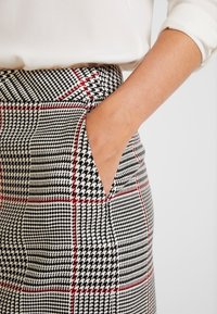 Jupe à motifs à carreaux noir et blanc avec accents rouges, présentant une silhouette élégante et une poche latérale, fabriquée en tissu texturé.