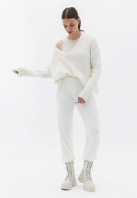 Witte geribbelde trui met een diepe V-hals en oversized pasvorm, gecombineerd met witte joggers met trekkoord en lichtgekleurde veterschoenen.