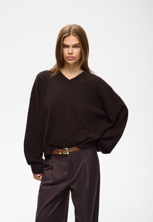 Jonge vrouw met stijl haar draagt een donkerbruine oversized trui, bruine broek en een slangenleren riem, staand tegen een effen witte achtergrond.