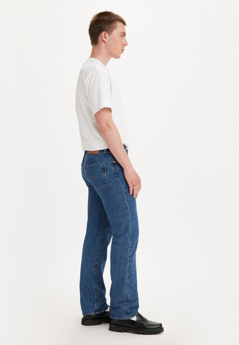 Un jeune homme de profil portant un t-shirt blanc, un jean bleu et des mocassins noirs sur un fond blanc uni.