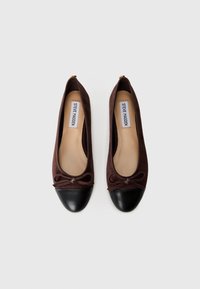Brown suede ballet flats with a black leather cap toe, featuring a bow detail at the front and a cushioned tan insole.