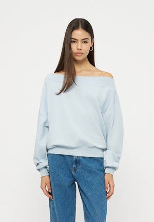 Femme aux cheveux longs et raides portant un sweat-shirt bleu clair à épaules dénudées et un jean bleu, debout devant un fond blanc uni.