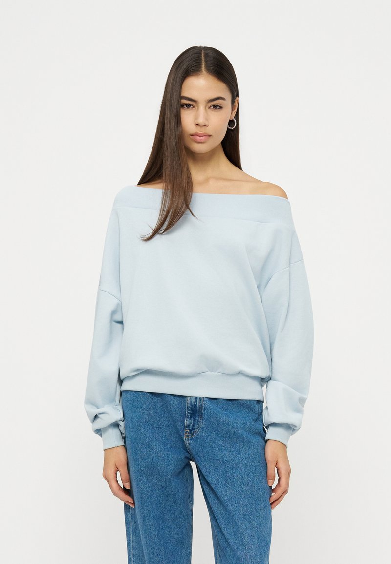 Femme aux cheveux longs et raides portant un sweat-shirt bleu clair à épaules dénudées et un jean bleu, debout devant un fond blanc uni.