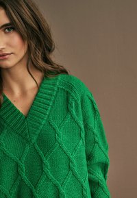 Maglione verde a maglia con scollatura a V e motivo a diamante testurizzato. Collo a costine e maniche ampie, realizzato in un filato spesso.