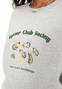 Sivá mikina s grafickým motívom ustrice a zeleným textom. Dizajn obsahuje škrupinu ustrice, plátok citrónu a misku so zelenou zeleninou.