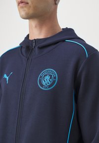 Námornícky zip-up mikina vyrobená z mäkkej tkaniny, s svetlomodrými akcentmi a okrúhlym logom Manchester City na hrudi.