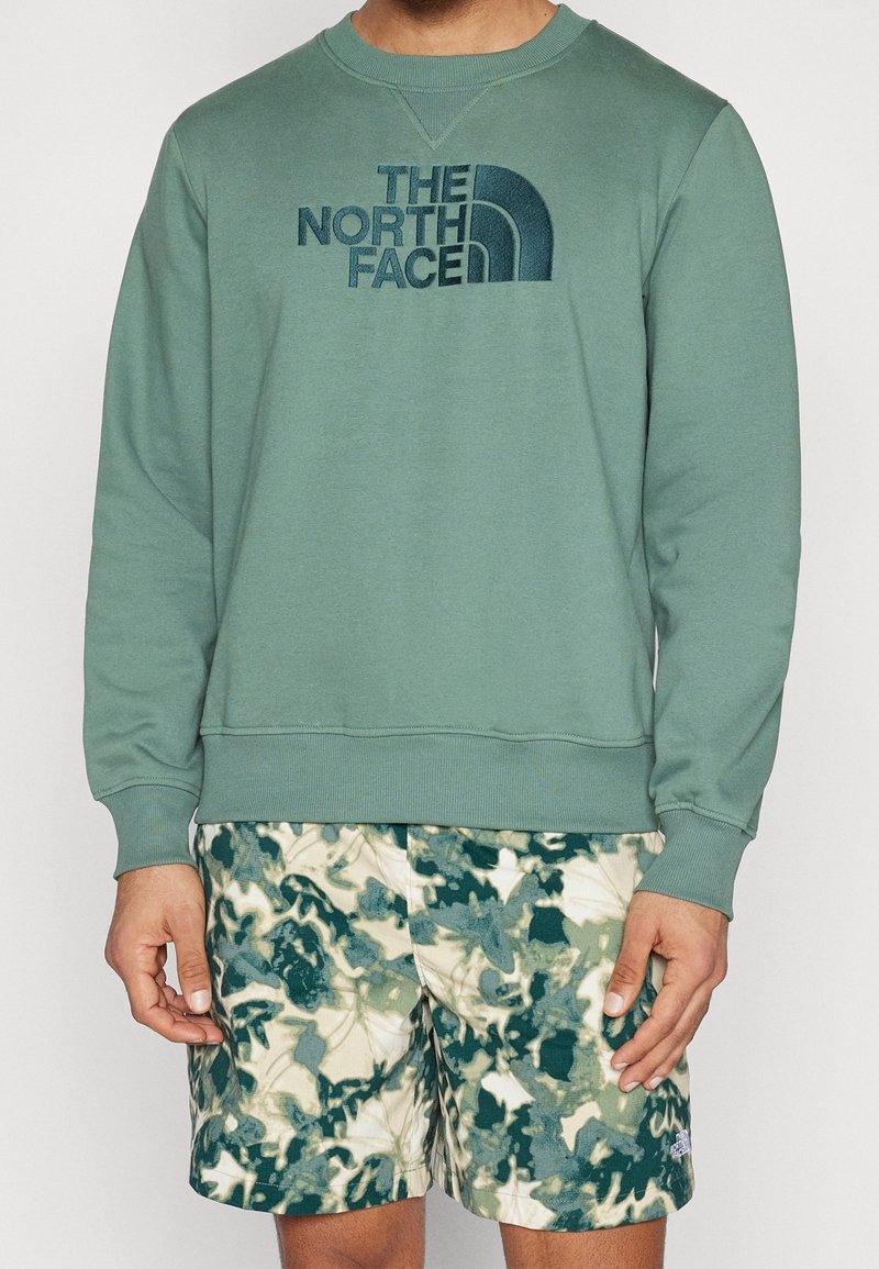 Homme portant un sweat-shirt The North Face vert clair et un short à motif camouflage vert et blanc.
