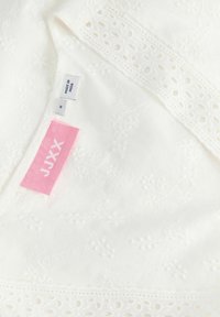Tissu blanc brodé avec bordure en dentelle, montrant l'étiquette de marque rose JJXX ainsi que les petites étiquettes de taille et fabriqué en Inde.
