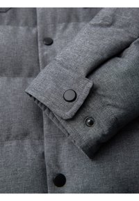 Gros plan d'une manche de veste matelassée grise avec des boutons-pression noirs et un tissu texturé.