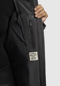 Must mustaga jope sisemus, kus on nähtaval tõmblukuga tasku ja kangast sildiga, millel on tekst. Sile tekstuur, kerge disain ja soe materjal.