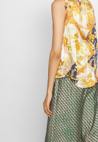 Farverig ærmeløs top med et blomstret og geometrisk mønster i gul og marineblå, parret med en plisseret grøn nederdel med et diamantmønster.