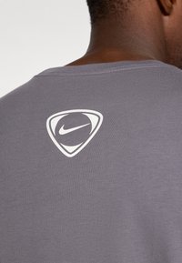 T-shirt grigio a maniche corte con collo rotondo, caratterizzato da un grande logo Nike bianco a forma di scudo sul retro. Texture liscia.