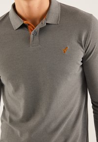 Harmaa pitkähihaisen polo-paidan, jossa on teksturoitu pinta. Paidassa on oranssi yksityiskohta kauluksen alla ja pieni oranssi brodeerattu logo rinnassa.
