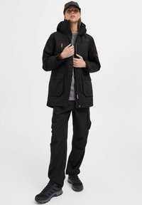 Schwarze wasserdichte Jacke mit Kapuze, ausgestattet mit mehreren Fronttaschen und einem Reißverschluss. Kombiniert mit schwarzen Cargo-Hosen und Wanderschuhen.