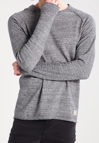 Grå långärmad stickad tröja med strukturerad tyg, raglanärmar och en liten logotypetikett längst ner till vänster. Svarta byxor är på.