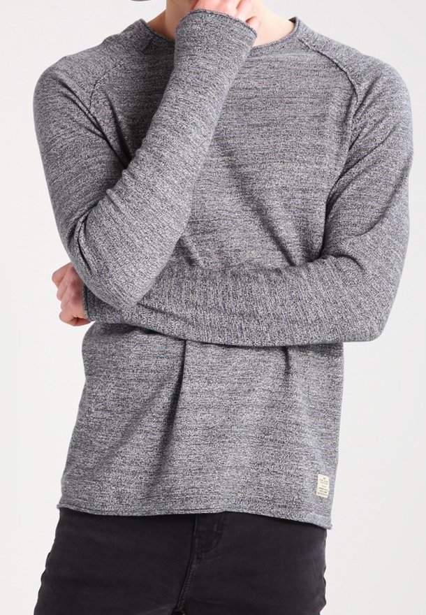Grå långärmad stickad tröja med strukturerad tyg, raglanärmar och en liten logotypetikett längst ner till vänster. Svarta byxor är på.