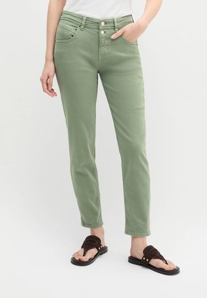 Persona che indossa pantaloni slim fit verde chiaro e sandali marroni, in piedi con una mano in tasca davanti a uno sfondo semplice.
