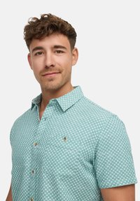 Korte mouwen, muntgroen shirt met een geometrisch wit patroon, een button-down kraag en een borstzak met een gouden knop.