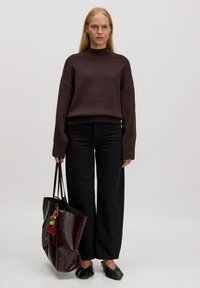 Dunkelbrauner gestrickter Pullover mit hohem Kragen, kombiniert mit schwarzen weit geschnittenen Hosen. Hält eine glänzende burgunderfarbene Tote-Tasche mit bunten Charms.