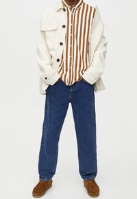 Personne portant une chemise rayée verticale marron et blanche, une veste boutonnée écrue, un jean bleu et des chaussures en daim marron, debout les mains dans les poches.