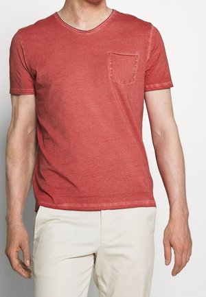 Red cotton t-shirt featuring short sleeves, a v-neckline, and a chest pocket. The fabric has a slightly worn texture with subtle color variations.