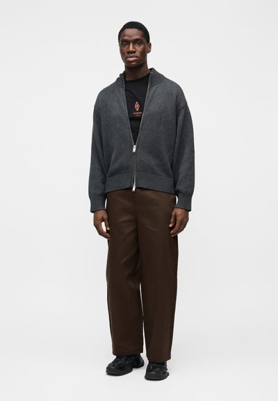Sivý pletený sveter na zips s voľným strihom, nosený cez čiernu košeľu s červeným logom. Kombinovaný s hnedými širokými nohavicami a čiernymi teniskami.