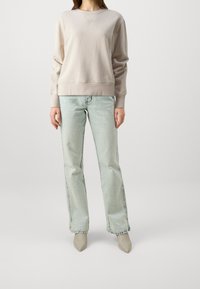 Vaaleanbeige ribbineulepaita pyöreällä kaula-aukolla ja kirjailulla logolla, yhdistettynä vaaleansinisiin suoralahkeisiin farkkuihin ja beigenvärisiin teräväkärkisiin korkokenkiin.